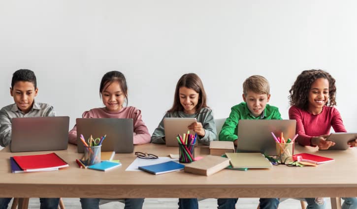 Five children sit at a wooden table, each using a laptop with books, notebooks, and colorful supplies in a bright classroom setting.